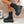 Black platform boots with multiple straps worn by a person against a disco ball and tiled wall background.
