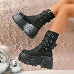 Black platform boots with buckles worn by a person, against a mosaic tile wall background.