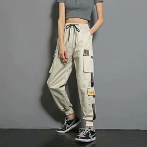 Khaki label techwear joggers with black and yellow patches on the cargo pockets, woman standing in three-quarter view