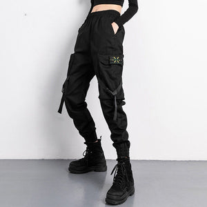 Black strap techwear joggers, front view with a hanging strap, green badge on the cargo pocket and black boots
