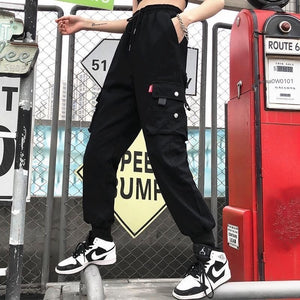 Black snap pocket techwear joggers worn on a city street, side view of the snap-flap cargo pocket and red tab