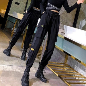 Two women in black label cargo joggers with yellow label patches, front view, tapered legs into black boots