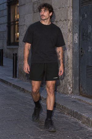 Man walking down a stone street in the Black Baggy Lined Gym Shorts and black tee, front view with logo