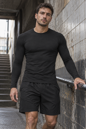 Model in the black compression shirt leaning on a railing in a tiled stairwell, front three-quarter view