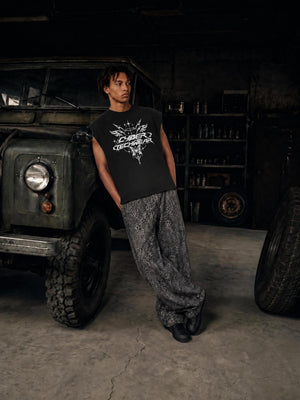 A model leans against a parked vehicle in a workshop wearing the black tank with wide printed trousers
