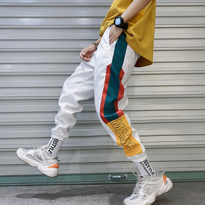 Hvide colorblock techwear-joggers set fra siden under gang, med petrolblå og røde striber og et gult panel med bogstaver