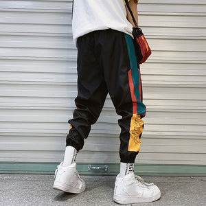 Sorte colorblock-joggers set bagfra, med petrolblå og røde sidestriber og et gult panel over hvide sneakers