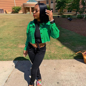 Green cropped denim jacket over a black top and black jeans, front view on a sunny campus walkway
