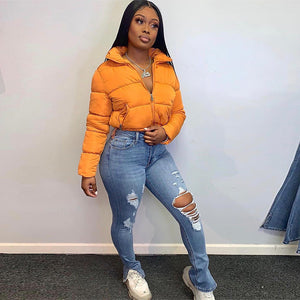 Kvinde i den orange Cropped Puffer Y2K-jakke og jeans med flænger, helfigur forfra mod en væg