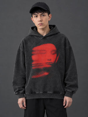 Man in a black cap wearing the Red Blur Face hoodie, front view of the blurred red portrait on washed black fleece
