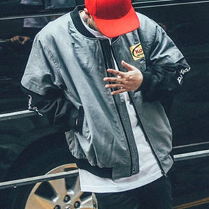 Man in the grey baseball bomber jacket with black sleeves next to a car, three-quarter front view