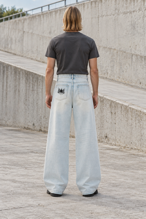 Back view of the light wash baggy jeans on a model, black spiked emblem on the back pocket, loose legs