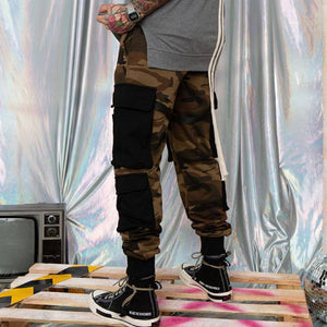 Camouflage cargo joggers worn by a model on a pallet, black stacked pockets on the thigh and ribbed black cuffs