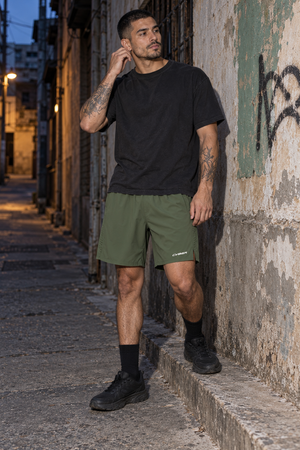 Model leaning on a wall at dusk in the Army Green nylon athletic shorts, front view with a black tee