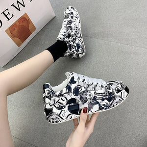 Hand holding a Black painted platform shoe next to a foot wearing the pair on a grey floor