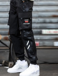 Black techwear cargo joggers in a side view with white sneakers, thigh pocket with red label and white shin stripes