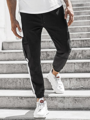Μαύρο jogger τεκγουέαρ με πλαϊνή ρίγα φορεμένο από άνδρα σε πέτρινα σκαλιά, μπροστινή όψη με τσέπες με καπάκι και λευκά sneakers