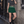 Model leaning on a stone wall in the Blackish Green pleated tennis skirt, front view with a grey crop tee