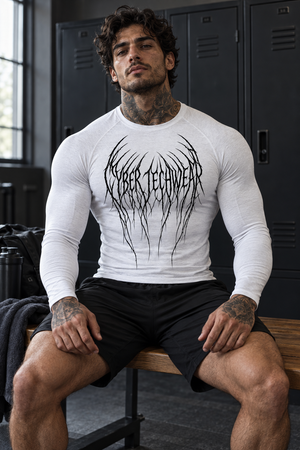 A tattooed man sits on a locker room bench in the white compression shirt, front view with shorts